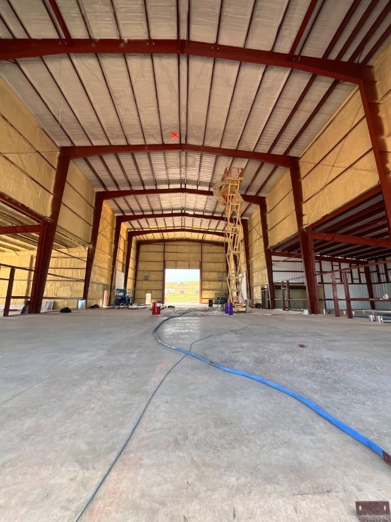 Vast interior of a large red-iron commercial shop north of Gillette, Wyoming, with cream/tan closed-cell spray foam covering the walls and roof deck between red roof purlins, scissor lift staged inside the bay during the Falcon Construction / MC Welding insulation project.