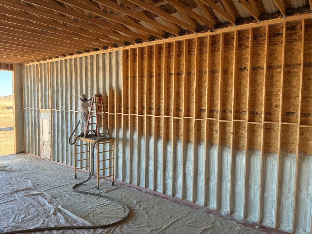 Seamless Systems crew member on a rolling scaffold inside a tall wood-framed hangar south of Buffalo, Wyoming, spraying closed-cell foam onto the upper wall studs with prairie visible through the open bay door.