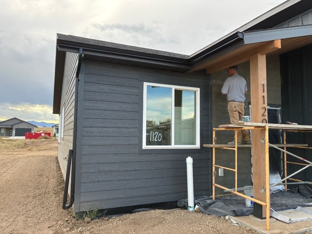 Seamless Systems crew member on a ladder finishing seamless black gutters along the front eave of a dark-grey new-build home at 1120 in a Sheridan, Wyoming housing development, with downspout already tied in at the porch corner.